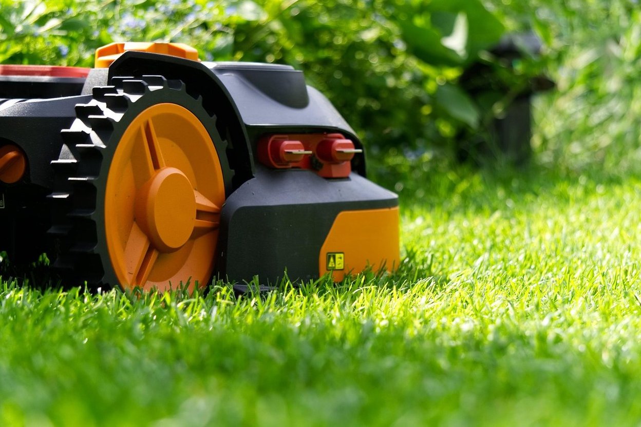 A robot mower cutting a flat, open lawn edge to edge beside a garden bed, with a homeowner trimming the grass along the border by hand nearby.