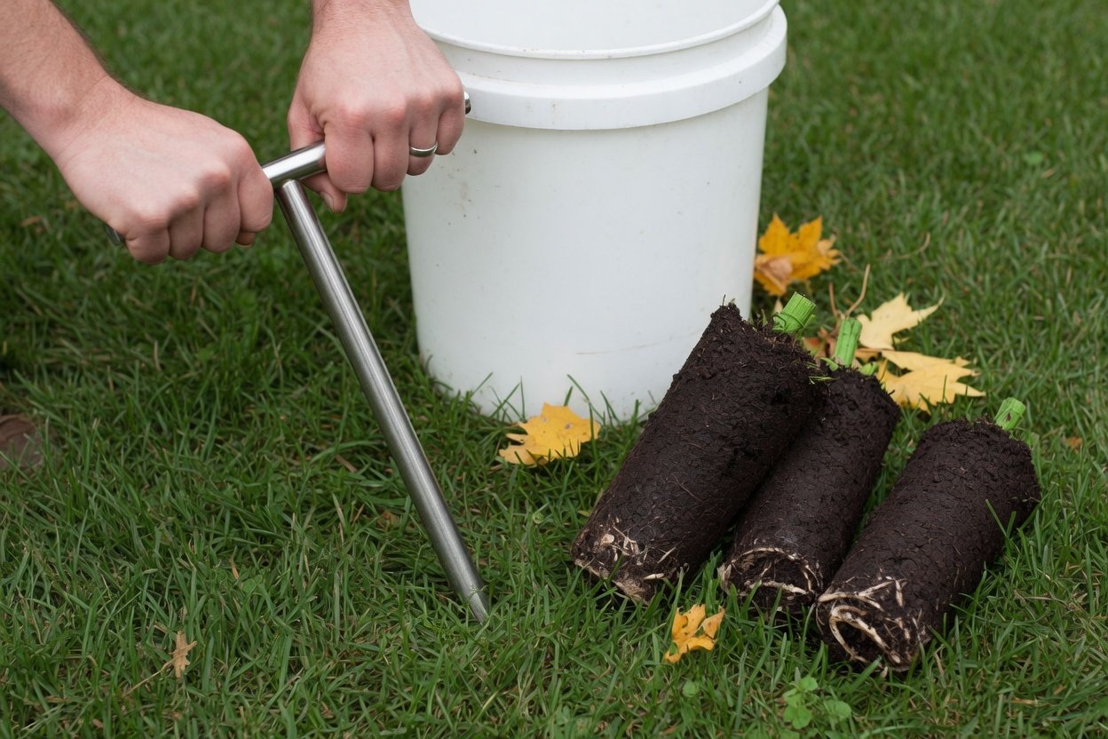 A soil probe pushed into a lawn beside soil sample cores and a bucket
