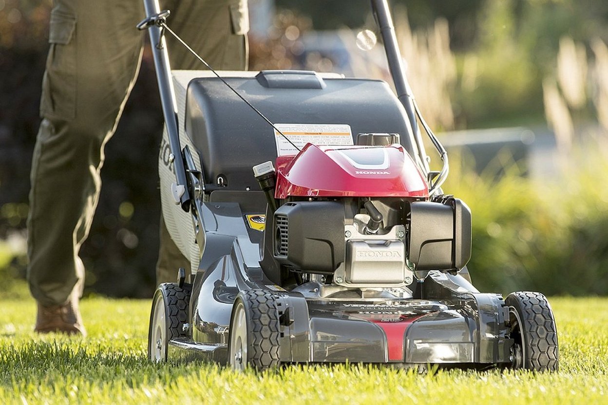 A homeowner pushing a battery rotary mower across a small, flat front lawn in late fall, with a garage door visible behind.